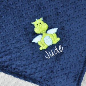 May include: Navy blue baby blanket with a textured, dotted pattern. A green and light blue dragon applique is centered on the blanket, with the name "Jude" embroidered below in white script. The blanket has a white, dotted backing.