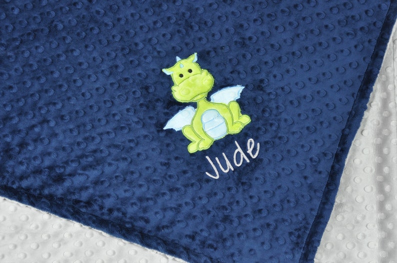 May include: Navy blue baby blanket with a textured, dotted pattern. A green and light blue dragon applique is centered on the blanket, with the name "Jude" embroidered below in white script. The blanket has a white, dotted backing.