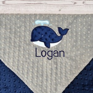 May include: A grey and navy blue baby blanket with a whale and the name "Logan" embroidered on it.