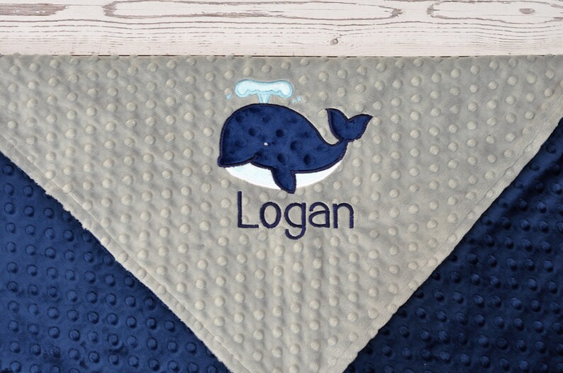 May include: A grey and navy blue baby blanket with a whale and the name "Logan" embroidered on it.