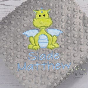 May include: A gray, textured baby blanket with a lime green and blue embroidered dragon design. The blanket has the name "Slade Matthew" embroidered in blue. The blanket is made of a soft, plush material.