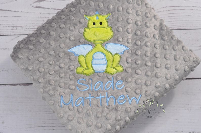 May include: A gray, textured baby blanket with a lime green and blue embroidered dragon design. The blanket has the name "Slade Matthew" embroidered in blue. The blanket is made of a soft, plush material.