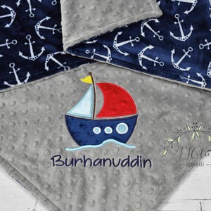 Puede incluir: Una manta para beb&eacute; azul marino y gris con tem&aacute;tica n&aacute;utica. La manta presenta anclas blancas sobre un fondo azul marino y un barco bordado con el nombre "Burhanuddin". La manta est&aacute; hecha de tela suave y texturizada.