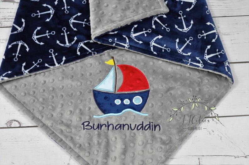 Puede incluir: Una manta para beb&eacute; azul marino y gris con tem&aacute;tica n&aacute;utica. La manta presenta anclas blancas sobre un fondo azul marino y un barco bordado con el nombre "Burhanuddin". La manta est&aacute; hecha de tela suave y texturizada.