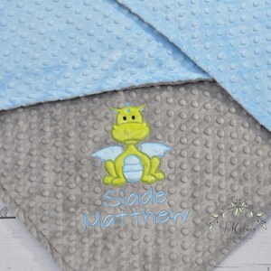 May include: A baby blanket with a light blue and gray color scheme. The blanket features a dragon applique in shades of yellow, green, and blue. The name "Slade Matthew" is embroidered in blue thread. The blanket has a textured, dotted pattern.