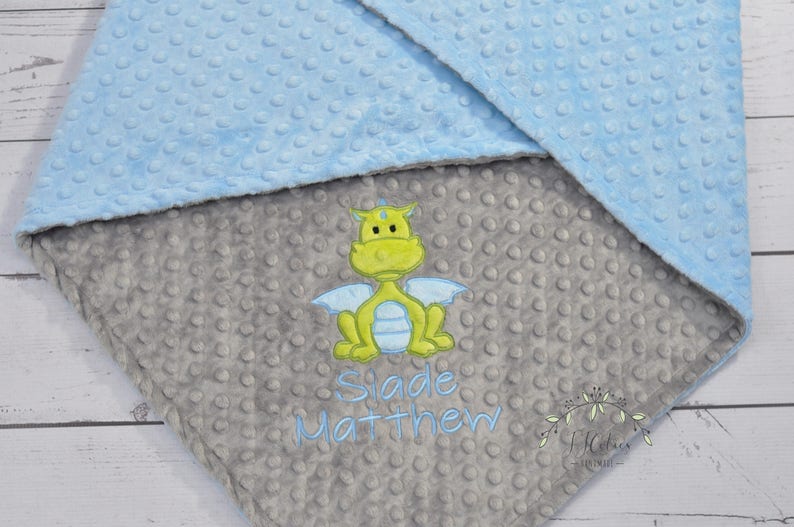 May include: A baby blanket with a light blue and gray color scheme. The blanket features a dragon applique in shades of yellow, green, and blue. The name "Slade Matthew" is embroidered in blue thread. The blanket has a textured, dotted pattern.