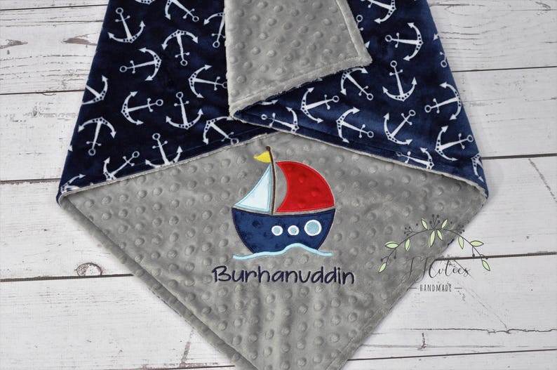 Puede incluir: Una manta para beb&eacute; azul marino y gris con tem&aacute;tica n&aacute;utica. La manta presenta patrones de anclas blancas sobre un fondo azul marino y un centro gris con un barco bordado y el nombre "Burhanuddin".