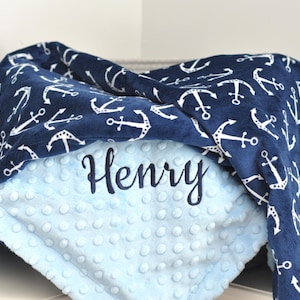 May include: A blue and white baby blanket with a navy blue side featuring a white anchor pattern and a light blue side with a textured dot pattern. The blanket is personalized with the name "Henry" embroidered in white.