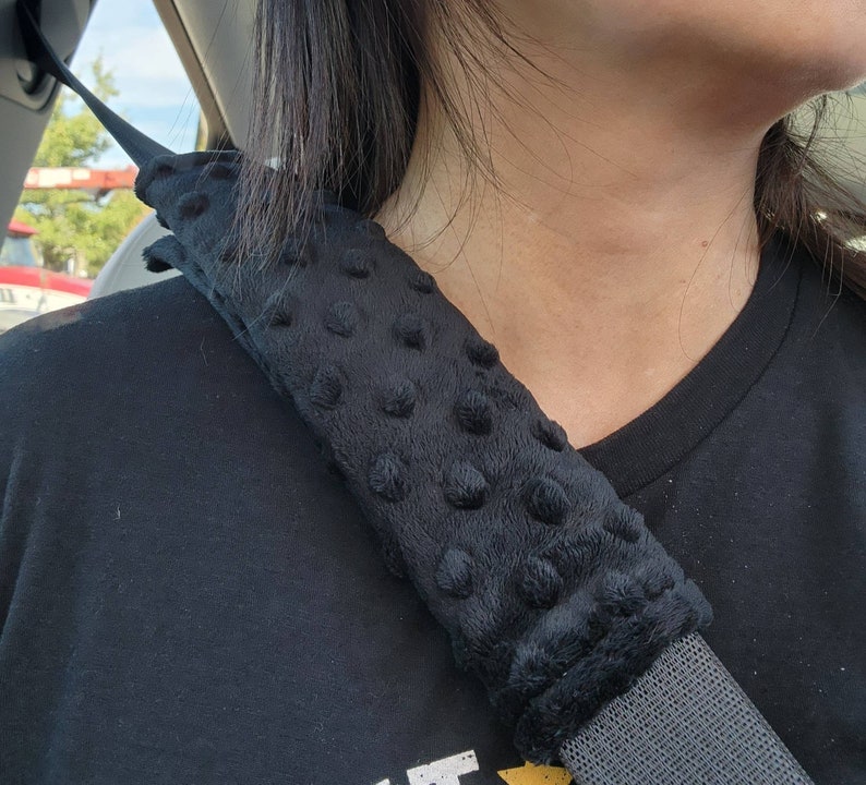 以下が含まれることがあります： 黒色の車のシートベルトカバーで、柔らかく、質感のある黒い生地で作られています。カバーには、隆起したドットパターンがあります。