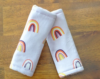 May include: Two purple fabric car seat belt covers with a rainbow pattern. The covers are soft and padded for comfort.