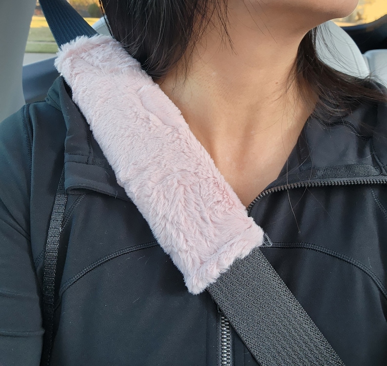 Puede incluir: Una funda para cintur&oacute;n de seguridad de coche de piel sint&eacute;tica rosa. La funda est&aacute; unida a un cintur&oacute;n de seguridad negro.