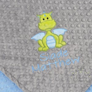 May include: A gray baby blanket with a textured, dotted surface. The blanket features an embroidered lime green dragon with light blue wings. The name "Slade Matthew" is embroidered in light blue. A light blue blanket edge is visible.