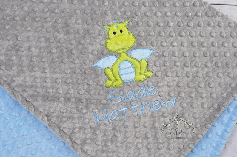 May include: A gray baby blanket with a textured, dotted surface. The blanket features an embroidered lime green dragon with light blue wings. The name "Slade Matthew" is embroidered in light blue. A light blue blanket edge is visible.