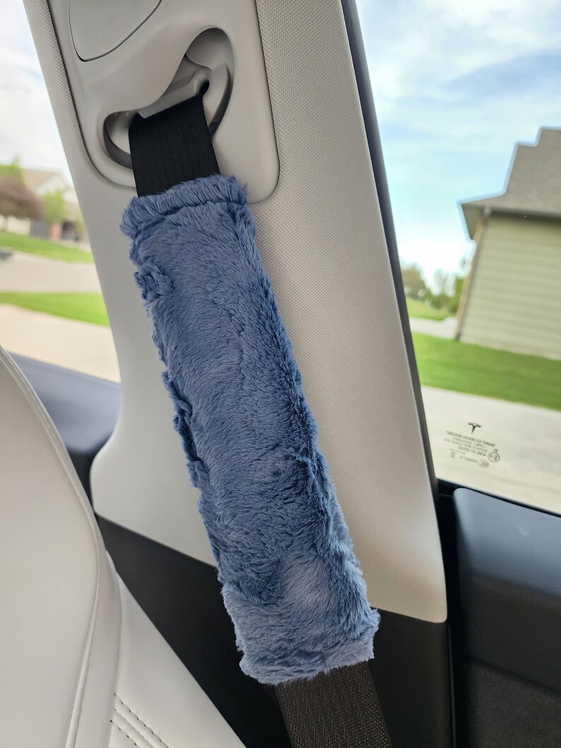 Puede incluir: Una funda de cintur&oacute;n de seguridad de coche de felpa azul. La funda est&aacute; unida a un cintur&oacute;n de seguridad negro y est&aacute; en un coche.