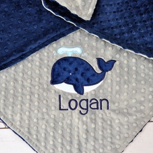 May include: A grey and navy blue baby blanket with a whale and the name "Logan" embroidered on it.
