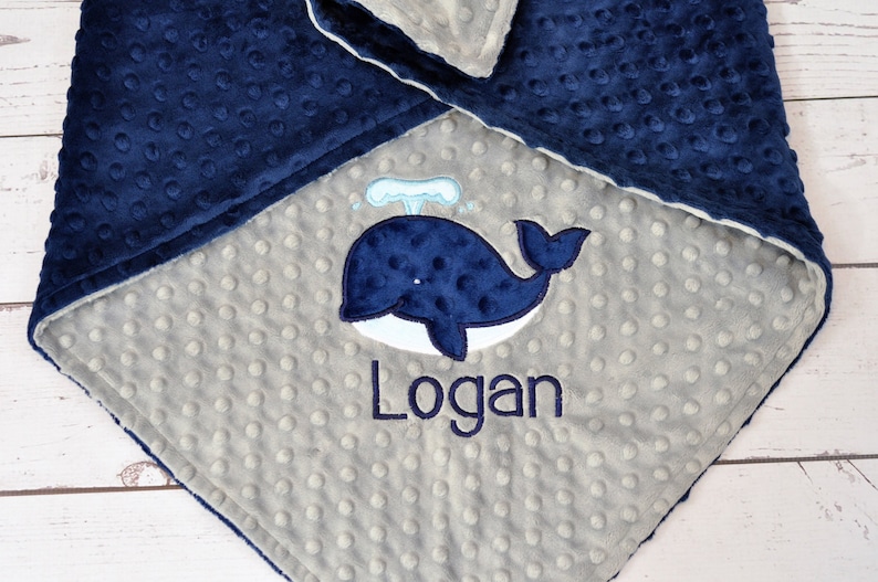 May include: A grey and navy blue baby blanket with a whale and the name "Logan" embroidered on it.