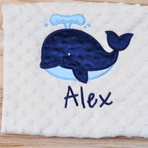 May include: A white baby blanket with a blue whale and water spout embroidered on it. The name "Alex" is embroidered below the whale.