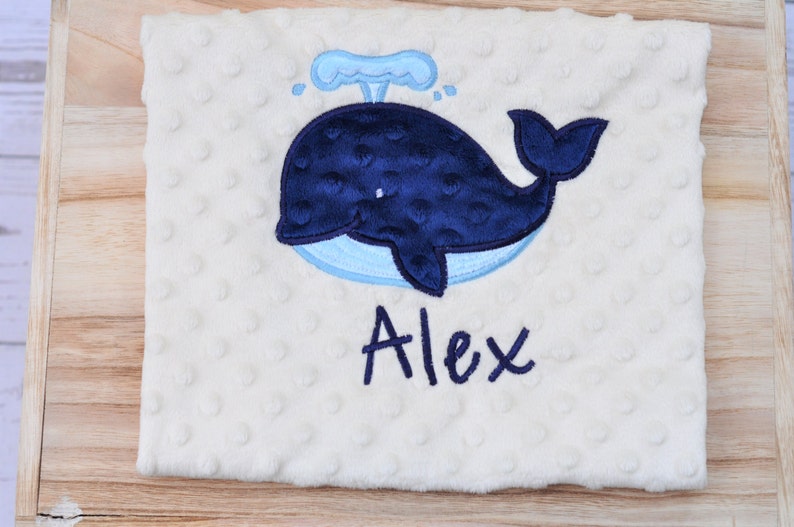 May include: A white baby blanket with a blue whale and water spout embroidered on it. The name "Alex" is embroidered below the whale.
