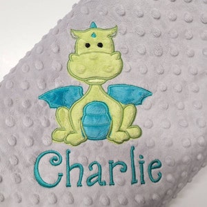 May include: A gray baby blanket with a textured, dotted surface. The blanket features an embroidered dragon in shades of green and blue. The name "Charlie" is embroidered in teal below the dragon.