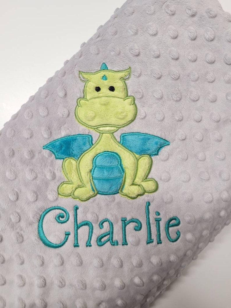 May include: A gray baby blanket with a textured, dotted surface. The blanket features an embroidered dragon in shades of green and blue. The name "Charlie" is embroidered in teal below the dragon.
