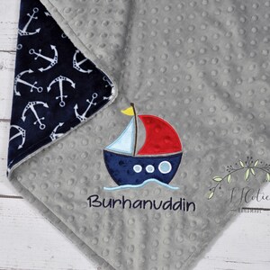 Puede incluir: Una manta gris para beb&eacute; con un borde estampado de anclas azul marino. La manta presenta un barco bordado con una vela roja y el nombre "Burhanuddin" en azul marino. La manta est&aacute; hecha de tela suave y texturizada.