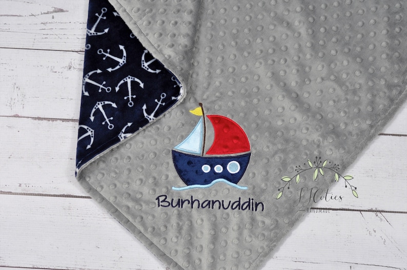 Puede incluir: Una manta gris para beb&eacute; con un borde estampado de anclas azul marino. La manta presenta un barco bordado con una vela roja y el nombre "Burhanuddin" en azul marino. La manta est&aacute; hecha de tela suave y texturizada.