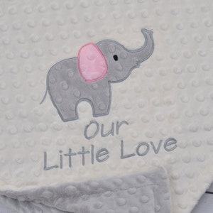 Puede incluir: Una manta de beb&eacute; blanca con un elefante bordado gris y rosa y el texto "Our Little Love".