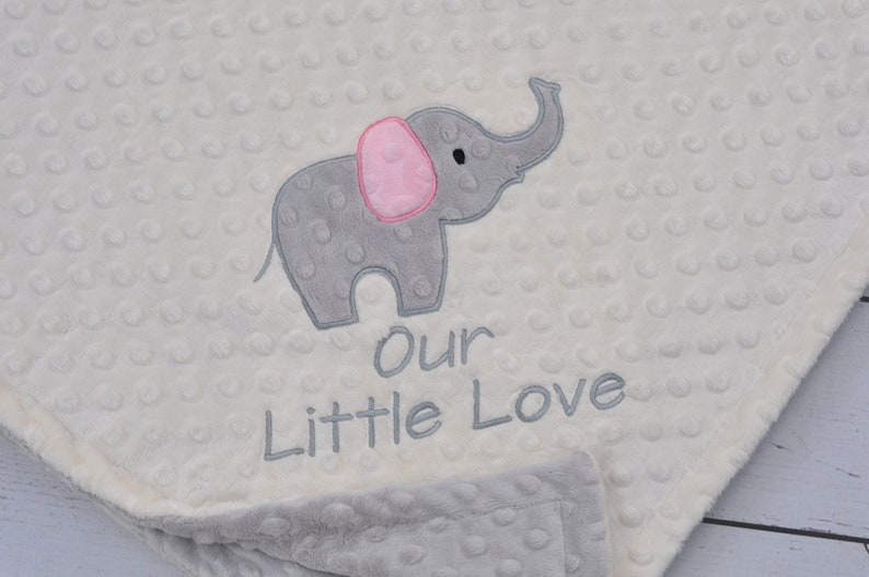 Puede incluir: Una manta de beb&eacute; blanca con un elefante bordado gris y rosa y el texto "Our Little Love".