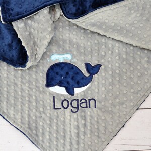 May include: A grey and navy blue baby blanket with a whale and the name "Logan" embroidered on it.