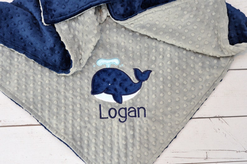 May include: A grey and navy blue baby blanket with a whale and the name "Logan" embroidered on it.