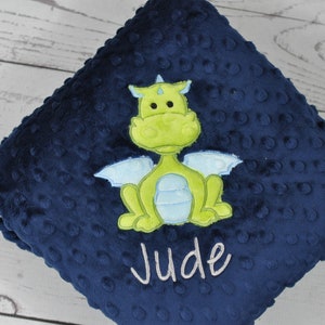 May include: A navy blue baby blanket with a textured surface and a lime green dragon applique. The dragon has light blue wings and is centered on the blanket. The name "Jude" is embroidered in white.