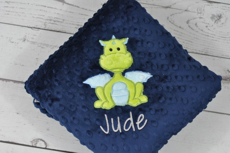 May include: A navy blue baby blanket with a textured surface and a lime green dragon applique. The dragon has light blue wings and is centered on the blanket. The name "Jude" is embroidered in white.