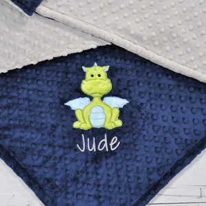 May include: A navy blue baby blanket with a green and blue dragon applique. The blanket has a textured, dotted pattern and is trimmed with a gray fabric. The name "Jude" is embroidered in white.