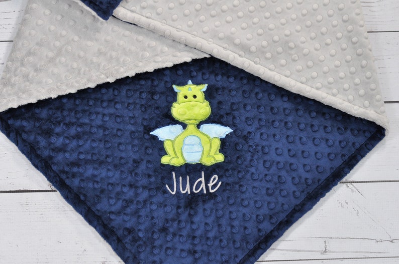May include: A navy blue baby blanket with a green and blue dragon applique. The blanket has a textured, dotted pattern and is trimmed with a gray fabric. The name "Jude" is embroidered in white.
