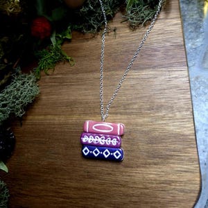 May include: A silver chain necklace with a charm of three stacked books. The books are painted in shades of pink, purple, and blue with white details.