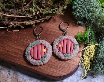 May include: A pair of earrings featuring a red door with a yellow knob, surrounded by a stone-like border and green accents. The earrings are made of polymer clay and have a hook closure.