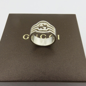 May include: A silver-toned ring featuring a cut-out design with the interlocking G logo at the center. The ring is positioned on a dark brown box with the gold-colored text GUCCI printed on the surface, clearly visible behind the metal band.