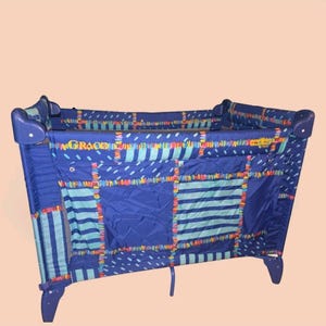 May include: A Graco portable playard featuring a blue fabric exterior with a multi-colored patchwork-style pattern of stripes, dashes, and dots. The rectangular structure has a mesh side panel and a yellow brand logo printed on the upper rail.