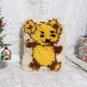 May include: A handmade, rectangular pillow featuring a teddy bear design. The bear is depicted in shades of yellow and brown against a cream-colored background. The pillow is made with a textured, looped yarn technique, giving it a cozy, vintage aesthetic.