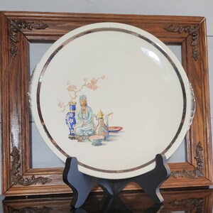 May include: A round off-white ceramic plate featuring a silver-toned rim and a central illustration of a seated figure, a blue and white vase, a floral-patterned teapot, and a small bowl, all positioned beneath a delicate pink flowering branch.