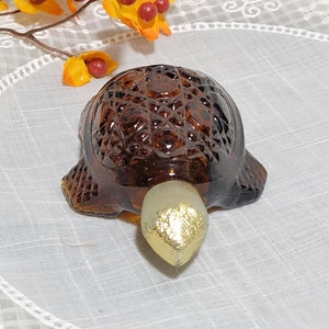 May include: A brown glass turtle figurine with a gold-colored head and shell. The turtle is sitting on a white lace tablecloth.