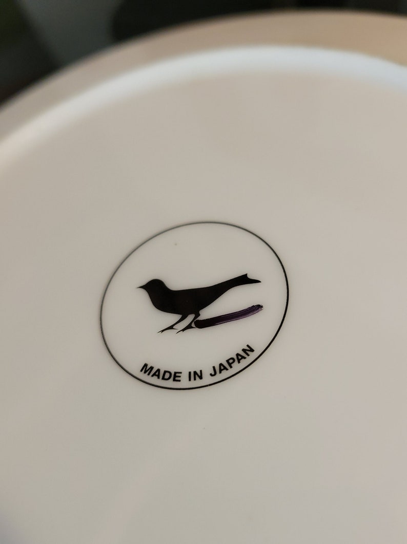 K&ouml;nnte beinhalten: Ein wei&szlig;er Keramikteller mit einer schwarzen Silhouette eines Vogels und dem Text "MADE IN JAPAN" in einem Kreis.