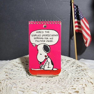 May include: A pink notepad with a spiral binding features a cartoon image of Snoopy writing with the text "Here's the world's greatest author working for his Pulitzer Prize..." and the signature "Schulz".