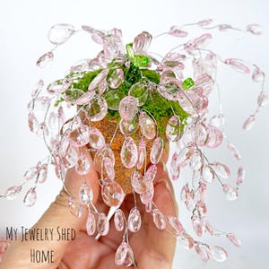 May include: A decorative miniature plant arrangement featuring pink crystal beads and green moss in a small, textured pot. The arrangement has delicate wire stems and cascading crystal droplets. The pot has a speckled, orange-brown finish.