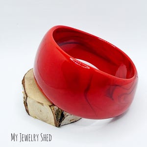 May include: A wide, glossy red bangle bracelet featuring subtle dark swirling patterns throughout its smooth, rounded surface. The bracelet is positioned at an angle, showcasing its thick, circular form and vibrant, solid red color profile.