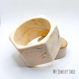 May include: Cream-colored, geometric bangle bracelet with a marbled pattern. The bracelet is a hexagonal shape and rests on a small wooden slice. The jewelry is from My Jewelry Shed.