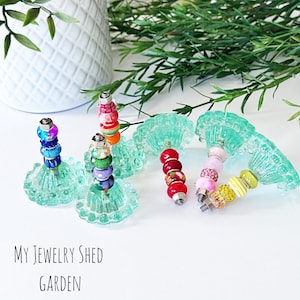 May include: Five colorful beaded jewelry pieces on turquoise stands. The stands are shaped like flower petals. The text "My Jewelry Shed Garden" is at the bottom of the image.
