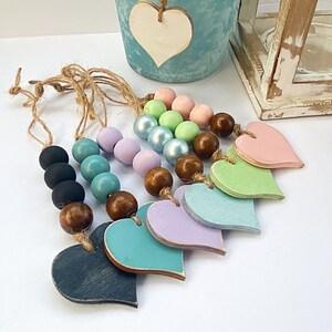 May include: A set of pastel colored wooden hearts with beaded strings. The beads are a mix of brown, light blue, and pastel colors. The hearts are in a rainbow order of colors, starting with pink and ending with teal.