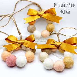 May include: Three felt ball wreaths in shades of pink, cream, tan, and gold, each adorned with a gold satin bow and twine for hanging. The text "My Jewelry Shed Holiday" is visible in the upper right corner.