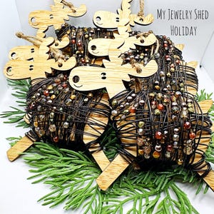 May include: Wooden moose ornaments with wire-wrapped bead bodies. The moose have smiling faces and are strung with twine for hanging. The beads are in shades of brown, gold, and red. The text "My Jewelry Shed Holiday" is in the upper right corner.